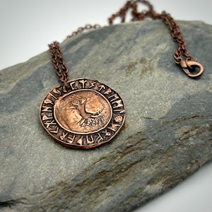 May include: A circular copper pendant necklace featuring an embossed animal figure in the centre surrounded by a border of etched runic characters. The pendant is suspended from a matching copper chain with a lobster claw clasp closure.