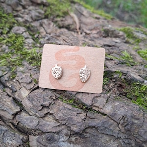 Puede incluir: Un par de pendientes de botón de fresa de color dorado sobre una tarjeta marrón con la palabra "S" impresa.