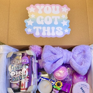 Op de afbeelding: Een geschenkdoos gevuld met paarse zelfzorgartikelen. De doos bevat een hoofdband, badzout, zeep, douchetabletten en een reisverpakking met lipgloss en andere schoonheidsproducten. De doos heeft ook een sticker met de tekst "YOU GOT THIS".