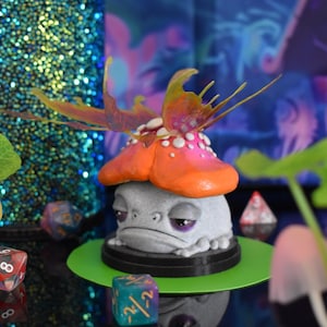 May include: A small, gray, frog-like creature with purple eyes is wearing a pink and orange mushroom hat with a iridescent butterfly wing on top. The creature is sitting on a green base with a black rim.