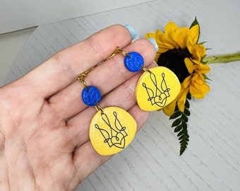 Puede incluir: Pendientes amarillos y azules con un diseño de tridente ucraniano. Los pendientes están hechos de arcilla polimérica y tienen un gancho de color dorado.