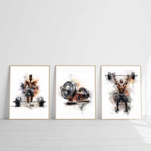 May include: A set of three framed art prints with watercolour-style illustrations of weightlifting. Each print shows a muscular figure with barbells, in shades of grey, black, and orange, against a white background.
