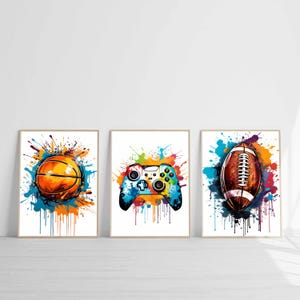 Könnte beinhalten: Set aus drei gerahmten Kunstdrucken mit Aquarellillustrationen. Enthält einen Basketball, einen Videospiel-Controller und einen Football, jeweils mit farbenfrohen Farbspritzern im Hintergrund. Die Drucke sind in Holzrahmen.