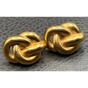 May include: A pair of gold-toned metal earrings featuring a knotted design. Each earring consists of a continuous, looped strand of metal that forms a rounded, intertwined shape with a smooth, polished surface and subtle metallic luster.
