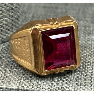 May include: A gold-toned ring featuring a large, rectangular, deep red gemstone. The ring has a textured design on the sides and a smooth, polished finish on the band. The gemstone is set in a square bezel.
