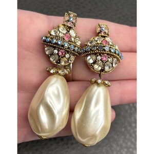 May include: A pair of drop earrings featuring a gold-tone orb design encrusted with clear, pink, and blue crystals. Each earring is topped with a small cross and finished with a large, smooth, teardrop-shaped cream-colored faux pearl pendant.