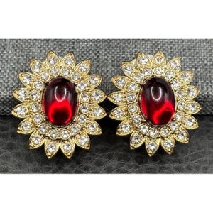 May include: A pair of gold-tone earrings featuring a large, oval-shaped deep red cabochon stone at the center. The stone is surrounded by a double layer of small, clear, round-cut rhinestones arranged in a sunburst or floral petal pattern.