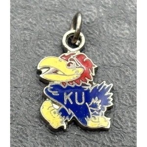May include: A silver-toned metal charm featuring an enamelled graphic of a bird mascot with a yellow beak, red hair, and a blue body. The blue section of the bird displays the white letters KU. The charm includes a small circular jump ring at the top.
