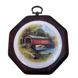 May include: An octagonal wall plaque featuring a dark wood frame and a central circular ceramic insert. The insert displays a colorful illustration of a red covered bridge over a river, surrounded by green trees, with a small boat near the bank.