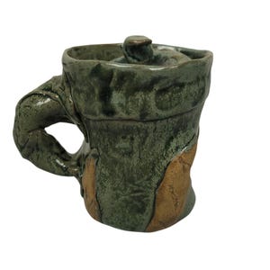 May include: Handmade ceramic mug with a lid, featuring a textured, organic design. The mug is primarily dark green with accents of brown and a sculpted handle. The lid has a small knob for easy removal. A unique piece for enjoying beverages.