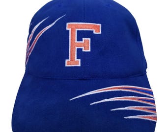 May include: A blue baseball cap with the letter "F" embroidered in orange. The cap features white and orange curved accents on the sides. The hat is a classic style with a curved brim.