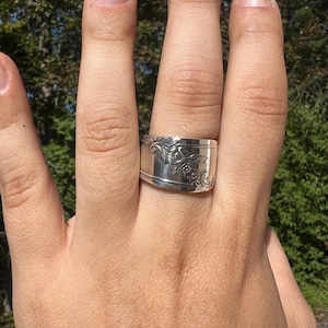 May include: A silver ring with floral and scrollwork designs. The ring is wide and has a slightly curved shape. The ring is worn on a finger, with a blurred green background.