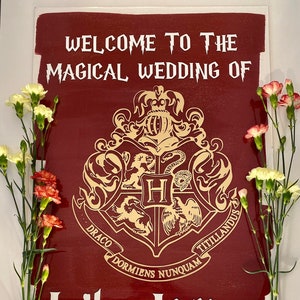 May include: A burgundy wooden sign with the words "WELCOME TO THE MAGICAL WEDDING OF" in white, along with a gold crest and the names "Lily & James". The sign is decorated with fresh flowers.