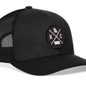 May include: Black trucker hat with a black and white embroidered patch featuring the letters "K" and "C" with a cross between them and the outline of the state of Missouri.