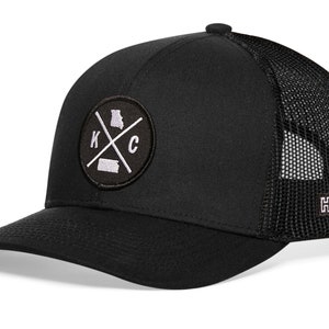 May include: Black trucker hat with a white embroidered patch featuring the letters "KC" with a cross and the outline of the state of Missouri. The hat has a black mesh back with a small black "H" logo.