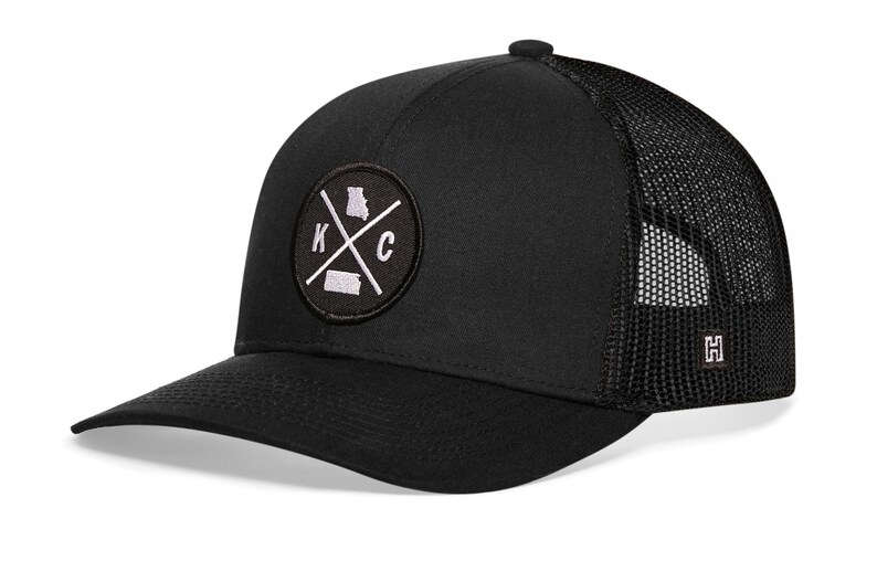 May include: Black trucker hat with a white embroidered patch featuring the letters "KC" with a cross and the outline of the state of Missouri. The hat has a black mesh back with a small black "H" logo.