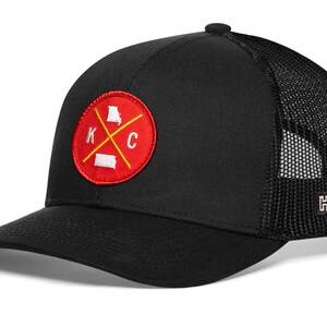 K&ouml;nnte beinhalten: Schwarze Baseballkappe mit Mesh-R&uuml;ckseite und gebogenem Schirm. Auf der Vorderseite befindet sich ein rotes rundes Patch mit wei&szlig;en Buchstaben "K" und "C" und gelbem Kreuz, mit Umrissen von Missouri und Kansas.