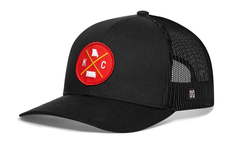 K&ouml;nnte beinhalten: Schwarze Baseballkappe mit Mesh-R&uuml;ckseite und gebogenem Schirm. Auf der Vorderseite befindet sich ein rotes rundes Patch mit wei&szlig;en Buchstaben "K" und "C" und gelbem Kreuz, mit Umrissen von Missouri und Kansas.