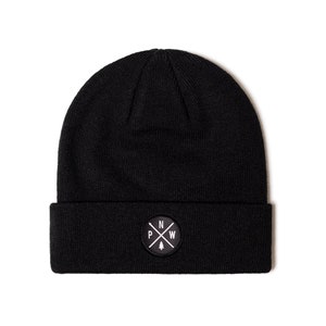 May include: Black beanie with a folded brim. A white circular patch on the front displays the letters "PNW" with a cross and a small evergreen tree. The beanie is made of a soft, knitted material.