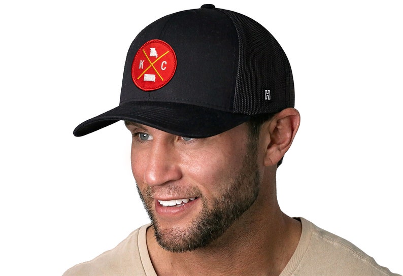 K&ouml;nnte beinhalten: Schwarze Trucker-Cap mit gebogenem Schirm und Mesh-R&uuml;ckseite. Auf der Vorderseite befindet sich ein kreisf&ouml;rmiger Aufn&auml;her mit rotem Hintergrund, den Buchstaben "K" und "C" sowie den Umrissen von Missouri und Kansas.