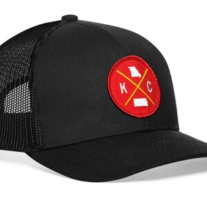 May include: Black trucker hat with a mesh back and a curved brim. The front features a red circular patch with a yellow cross and the letters "K" and "C", along with outlines of the states of Missouri and Kansas.