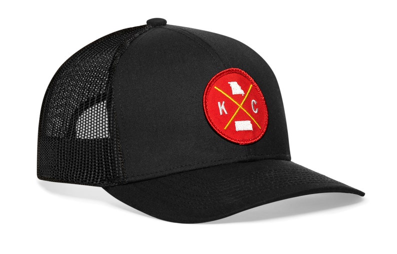 K&ouml;nnte beinhalten: Schwarze Trucker-Cap mit Mesh-R&uuml;ckseite und gebogenem Schirm. Auf der Vorderseite befindet sich ein roter runder Aufn&auml;her mit einem gelben Kreuz und den Buchstaben "K" und "C" sowie Umrissen von Missouri und Kansas.