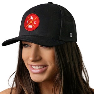 K&ouml;nnte beinhalten: Schwarze Baseballkappe mit Mesh-R&uuml;ckseite und gebogenem Schirm. Die Kappe hat einen runden Aufn&auml;her mit rotem Hintergrund, den Buchstaben "KC" und Umrissen der Bundesstaaten Kansas und Missouri. Ein kleines Metallschild befindet sich an der Seite.