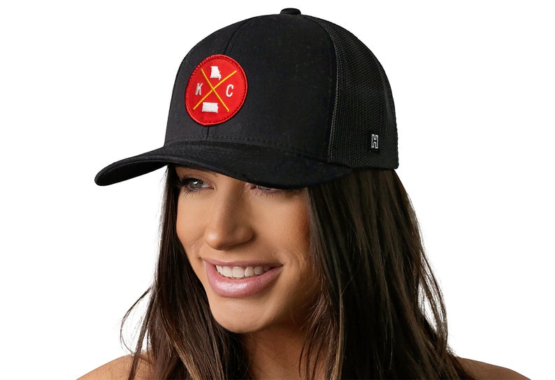 K&ouml;nnte beinhalten: Schwarze Baseballkappe mit Mesh-R&uuml;ckseite und gebogenem Schirm. Die Kappe hat einen runden Aufn&auml;her mit rotem Hintergrund, den Buchstaben "KC" und Umrissen der Bundesstaaten Kansas und Missouri. Ein kleines Metallschild befindet sich an der Seite.