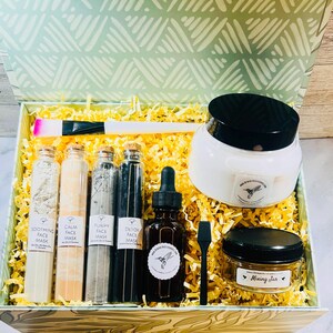 May include: A gift box filled with skincare products, including face masks, a serum, and a mixing jar. The box contains a white jar with a black lid, several glass bottles, and a brush. The box is lined with yellow paper and has a green and white patterned lid.