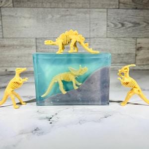 May include: Three yellow plastic dinosaur skeletons, one on top of a blue and purple soap bar, and two on the table. The soap bar has a blue and purple swirl design.