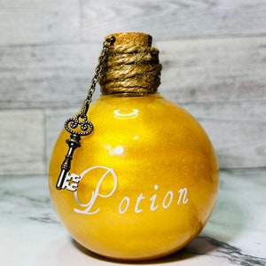 May include: A gold glass bottle with a cork stopper and a key charm. The bottle is filled with a glittery liquid and has the word "Potion" written on it in white.