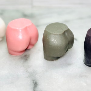 May include: Four small, butt-shaped objects in a row. The colors are white, pink, gray, and dark purple. The objects are smooth and appear to be made of a solid material. They are arranged on a white and gray marble surface.