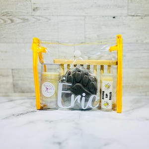 May include: A clear plastic bag with a yellow zipper contains a small gift set. The set includes a paw-shaped soap, a small jar of balm, and a lip balm tube. The lip balm tube has a bee design and the text "BOOM LIP BALM" on it. The paw-shaped soap has the name "Eric" written on it.
