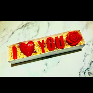 May include: A rectangular gift box contains red wax letters spelling "I LOVE YOU", a red heart, and a red rose. The box is filled with shredded yellow paper and sits on a white and gray marble surface.