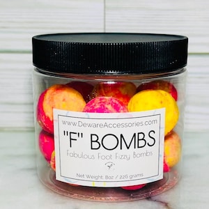 May include: A clear plastic jar with a black lid containing multiple round, multi-colored foot fizzy bombs in shades of pink, orange, and yellow. The white label reads www.DewareAccessories.com, "F" BOMBS, Fabulous Foot Fizzy Bombs, Net Weight 8oz.