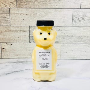 May include: A bear-shaped bottle filled with yellow bubble bath. The bottle has a black cap and a label that reads "Bubble Bear Bubble Bath." The background is a light wood pattern.