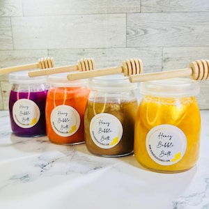 May include: Four glass jars filled with purple, orange, brown, and yellow liquid, each topped with a light-colored wooden honey dipper. Each jar features a circular white label with the text Honey Bubble Bath and a small yellow honey dipper icon.