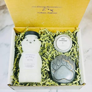 May include: A gift box filled with green shredded paper contains a white bottle with a bear-shaped design, a small round tin labeled "Mini Lotion Bar", and a gray paw-shaped soap.