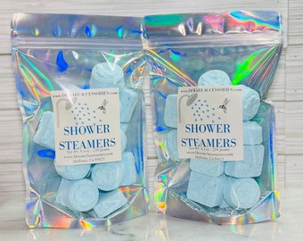 May include: Two clear plastic bags with blue shower steamers. Each bag contains eight blue shower steamers and has a white label with the text "Shower Steamers" and the company's website and address.