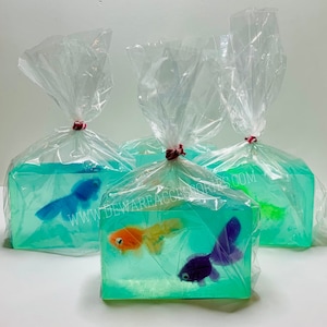 May include: Three clear plastic bags containing handmade soap bars shaped like fish tanks. Each soap bar features a different colored plastic fish inside. The soap bars are a light blue-green color. The bags are tied with red string. The text "www.dewareaccessories.com" is printed on the bag in the center.