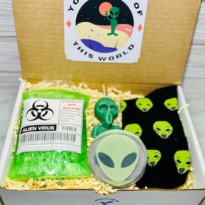 May include: A gift box filled with alien-themed items, including a green alien figurine, a green alien soap, a green alien virus-themed soap, and black socks with green alien faces. The box lid has a graphic of a green alien waving with the text "You're out of this world."