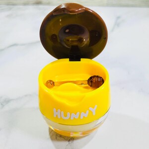 May include: A yellow honey pot with a brown lid that is open. The word HUNNY is printed in white letters on the front of the yellow container. A small brown honey dipper is resting horizontally inside the open pot. The base is clear plastic.