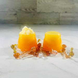 May include: Two small, orange jars, one filled with a light-colored, textured substance, the other with a yellow lid. Clear and amber-colored honey dippers surround the jars, suggesting a honey-themed product.