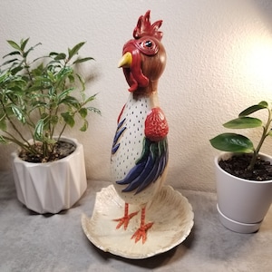 May include: A decorative rooster figurine standing on a textured, off-white circular base. The rooster has a brown head, a bright red comb and wattle, a yellow beak, and a white body with black speckles, red wings, and blue tail feathers.
