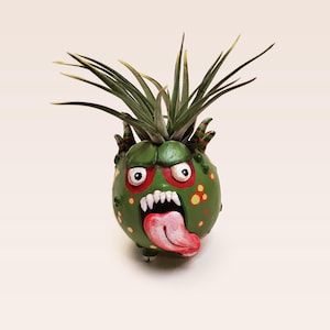 May include: A green, ceramic planter shaped like a monster with a toothy grin, red eyes, and a long pink tongue sticking out. The planter is decorated with yellow and orange spots and has a small air plant growing out of the top.
