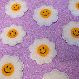 Puede incluir: Parches blancos con forma de margarita con un centro amarillo y una cara sonriente negra. Los parches están dispersos sobre una tela morada texturizada. Los parches son probablemente para fines decorativos.