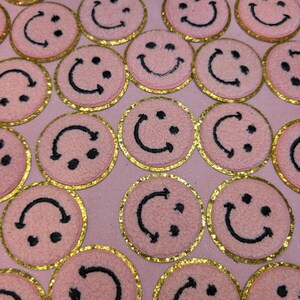 May include: A collection of pink and gold smiley face patches. The patches are made of a fuzzy material and have a gold glitter border.