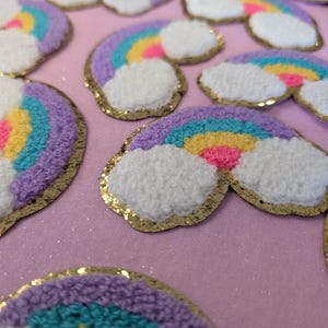 May include: A close-up of several rainbow-coloured felt patches with a fluffy white cloud design. The patches are outlined in gold glitter and are arranged on a pink sparkly surface.