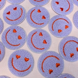 Puede incluir: Una colección de parches bordados con forma de cara sonriente con corazones por ojos, de color morado y naranja. Los parches están dispuestos en un patrón circular sobre un fondo rosa.