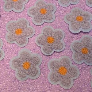 May include: A collection of flower-shaped patches. Each patch features a lavender-coloured, textured flower with an orange centre, outlined in light blue. The patches are scattered on a pink, patterned fabric.